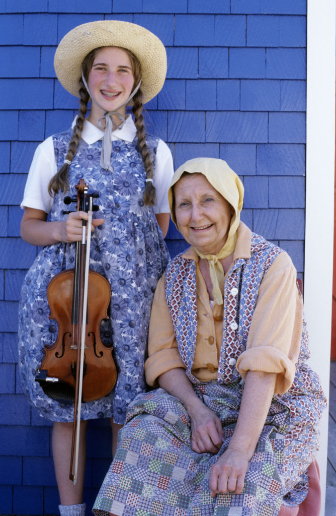 A young women with brown hair in pigtails, wearing a blue dress with a white collared undershirt, and holding a violin standing next to an older woman wearing a bonnet, muted yellow undershirt, and purple dress. 