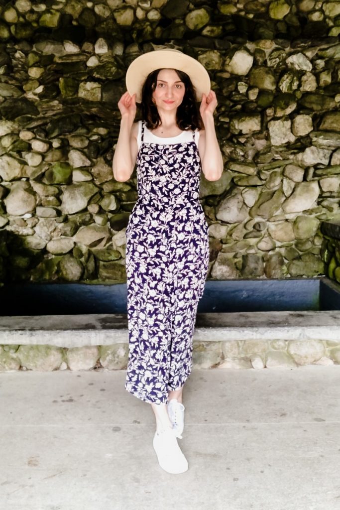 Dark-haired woman with light skin wearing a wide-brimmed hat and blue jumpsuit with white flowers and white sneakers.