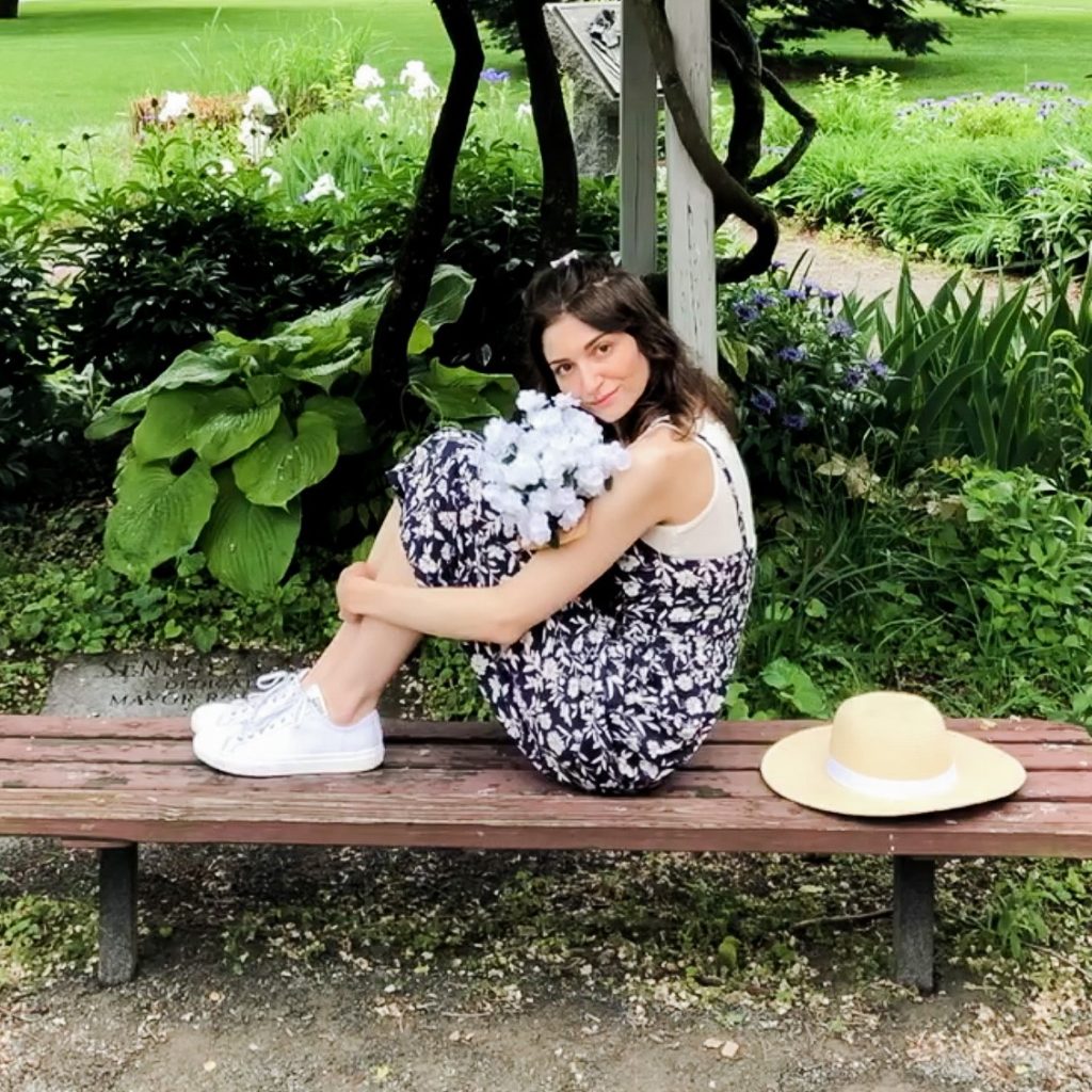 Dark-haired woman with light skin wearing a blue jumpsuit with white flowers and white sneakers.