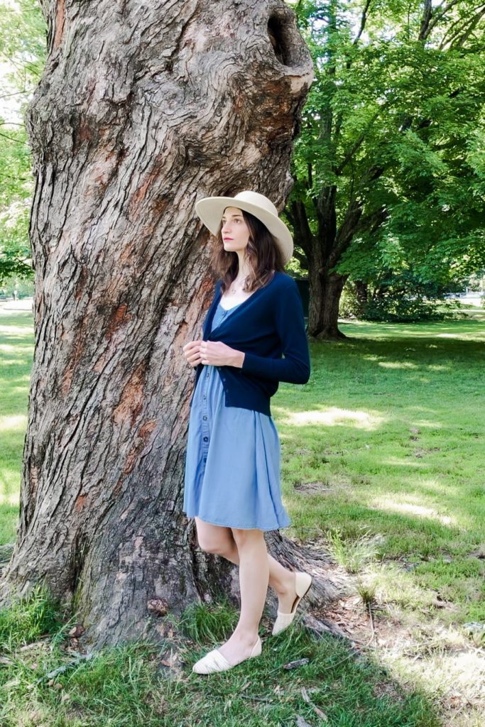 Dark-haired woman with light skin wearing a wide-brimmed hat, light blue dress, and a dark blue cardigan standing in front of a large tree.