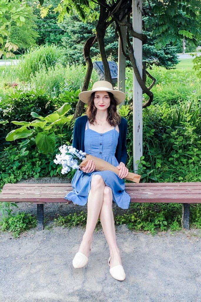 Dark-haired woman with light skin wearing a wide-brimmed hat, light blue dress, dark blue cardigan, and holding white flowers sitting on a bench in a garden.