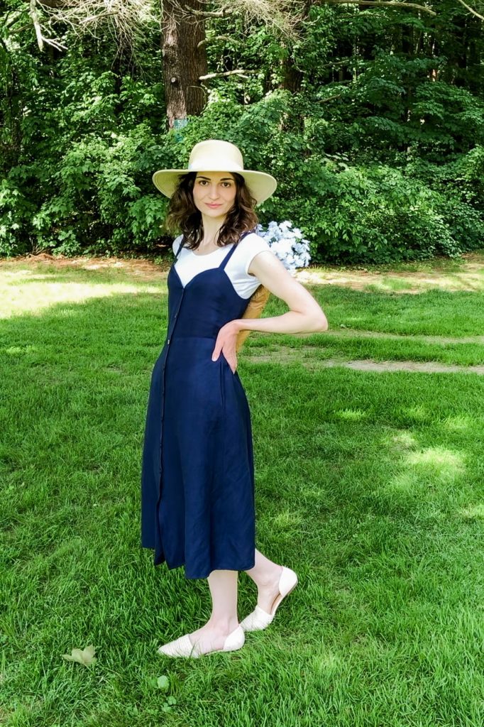 Dark-haired woman wearing a wide-brimmed hat, a long navy blue dress with a white shirt underneath, and flats holding a bouquet of white flowers.