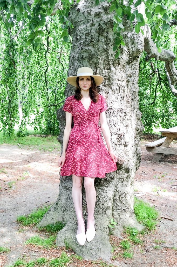 Dark-haired woman wearing a red floral dress and wide-brimmed hat standing in front of a tree.