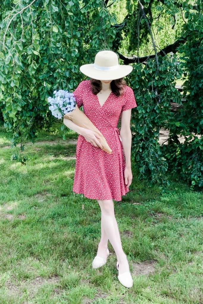 Dark-haired woman in a red floral dress hiding her face with a wide-brimmed hat and holding a bouquet of white flowers.